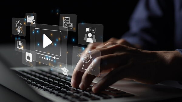 Hände tippen auf einem Laptop. Darüber schweben digitale Icons für Online-Kurse, Video und künstliche Intelligenz.